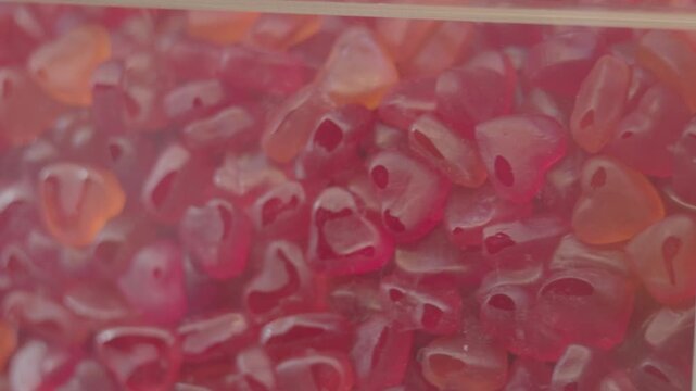 Close-up view of red jelly candy pieces in a transparent bin, revealing their soft, chewy texture and irregular shapes. The candies reflect ambient lighting and are densely packed, creating