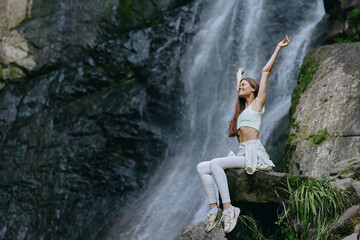 Young woman enjoying nature near waterfall, wearing white activewear, sitting on rock with arms...