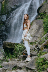 smiling woman enjoying nature at waterfall, wearing light sportswear and sneakers, surrounded by...
