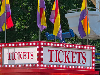 Brightly colored flags adorn the top of a ticket booth at an outdoor fair, with bold red lettering on the sign