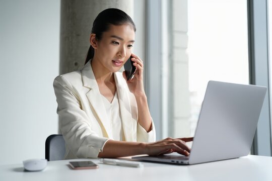 Asian business woman working on laptop and talking on phone in modern office space - Powered by Adobe
