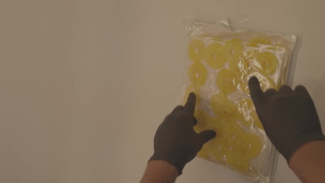 A gloved hand holds a vacuum-sealed transparent bag filled with perfectly shaped yellow dried pineapple rings. The background is a clean, white wall, and the focus is on the packaging