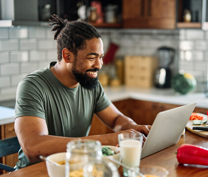 Happy young afro american man having fun preparing food and looking for recipes online using a laptop in kitchen, or a young businessman working from home office