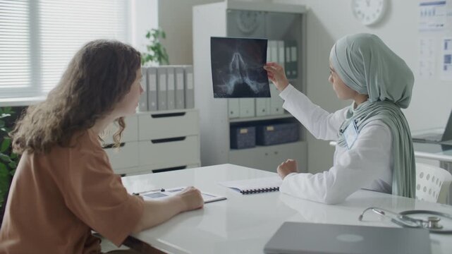 Female doctor in hijab and lab coat holding up X-ray film and discussing details with patient during consultation in medical office