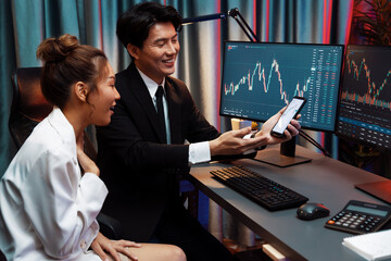 Asian businessman showing stock market application on mobile phone to partner pointing by pen,...