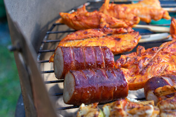 Close-up of grilled sausages and marinated chicken fillets cooking on an outdoor charcoal barbecue grill.