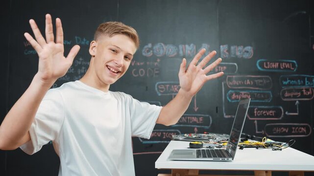 Side view of young boy programming code while looking at camera and greeting. high school student coding system while sitting at STEM classroom with electronic equipment scatter on table. Edification