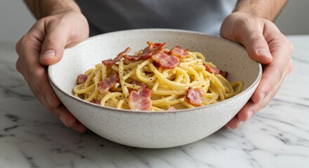 A person holding a bowl of carbonara pasta with bacon on a marble surface in a kitchen setting