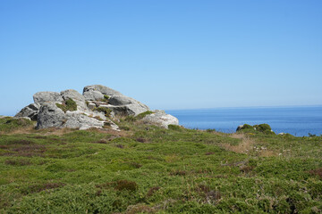 A weathered granite rock formation sits amidst a green and yellow heathland under a blue sky in Brittany, France.