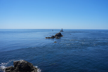 A distant view of a lighthouse and surrounding rocks in the sea, seen from the coast of Brittany, France.