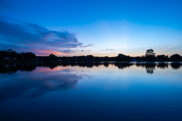 Fototapeta premium Twilight Over Lake with Soft Blue and Pink Reflections. A peaceful twilight scene unfolds over a calm lake, with soft hues of blue and pink painted across the sky and mirrored in the water. 