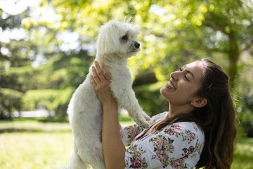 Smiling woman lifting her small white maltese dog in a green park, enjoying a sunny day