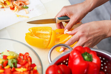 Hands cutting yellow bell pepper. Fresh vegetables on kitchen counter. Healthy food preparation at home. Red peppers in colander. Colorful salad ingredients. Woman cooking in domestic kitchen.