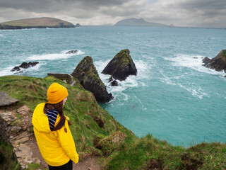 Girl tourist in yellow jacket and hat explore stunning Dunquin Pier area in county Kerry, Ireland....
