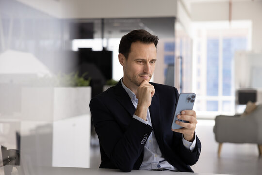 Businessman sit at desk in office using smartphone, checking social media for business-related updates or marketing, set reminders for tasks and deadlines, access or edit documents using mobile app - Powered by Adobe