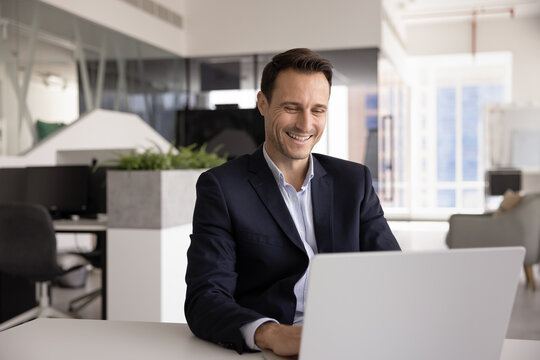 Middle-aged businessman sitting at table looks at his laptop with contented smile, receiving good news, completing task successfully, or enjoying his workflow in modern office. Tech usage in business