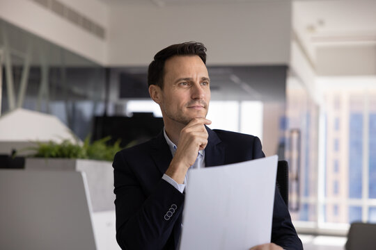 Focused professional man sits at his desk with open laptop, holding documents, gazes thoughtfully into distance with dreamy expression, engaged in reflection, planning, consider written information