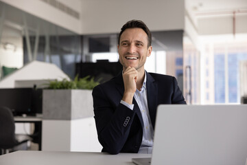 Positive middle-aged 40s man sits in workplace with laptop, distracted from workflow, looks away and laughing, deep in pleasant thoughts about career, future achievements, professional opportunities