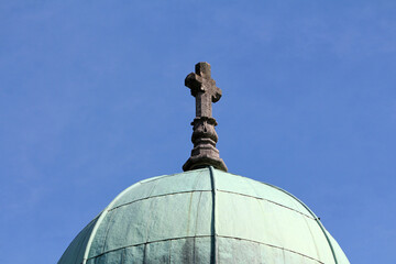 A weathered stone cross crowns the oxidized green copper dome of a historic building, set against a...