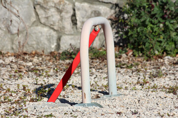 A sturdy, U-shaped metal parking barrier anchored with bolts and a red locking arm controls vehicle access on a gravel area beside a stone wall and green foliage