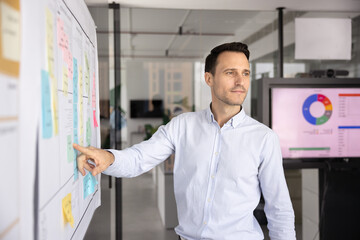 Pensive man looks aside, stand next to white board with attached colorful sticky notes with...