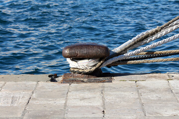 A robust metal mooring bollard tightly fastens thick ship ropes on a stone harbor quay, with rippling blue water providing a nautical backdrop in bright natural daylight