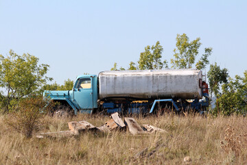 A faded blue water tanker truck sits unused in an overgrown grassy field, with patches of rust and weather stains on the metal tank and lush green trees in the background