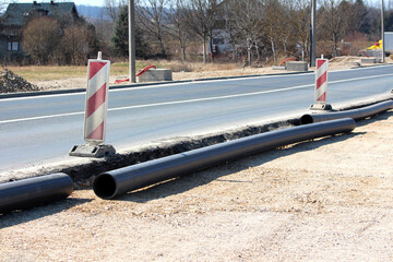 Black plastic utility pipes are laid out along a road under maintenance, marked by striped hazard...