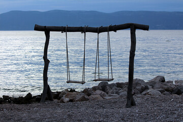 A handmade double swingset built from rough logs stands on a rocky lakeshore, silhouetted against...
