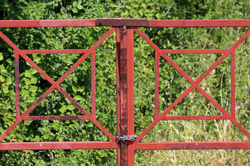 A weathered red metal gate features square geometric lattice patterns, speckled rust, and is...