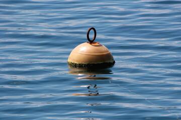 A semi-spherical, sun-faded mooring buoy with a rusty metal ring floats on gentle ripples, partially covered in green algae, serving as a maritime marker in open water