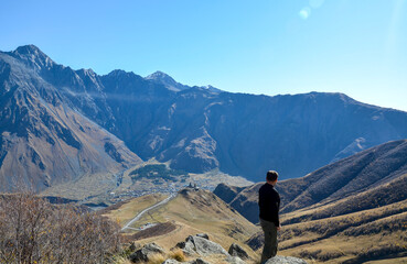 Solitary figure stands on a rocky outcrop, gazing at the Gergeti Trinity Church, perched dramatically above the village of Stepantsminda in the vast, mountainous landscape of Georgia