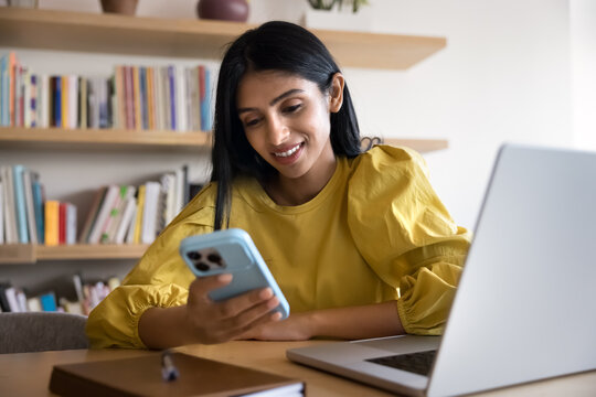 Young 25s Indian businesswoman holding smartphone using work-related mobile apps while sit at desk with laptop in modern office or library, managing schedule, tracking task, or accessing digital tool