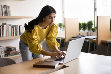Focused Indian office employee typing on laptop keyboard standing leaning at desk, reviewing received information or document, e-mailing to client, researching, check updates, working on presentation