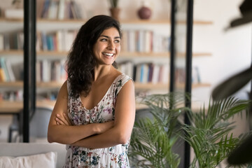 Confident Indian woman standing in professional workspace looks aside with smile, exudes positivity and self-assurance, enjoy workday results, thinks on future business opportunities or career growth