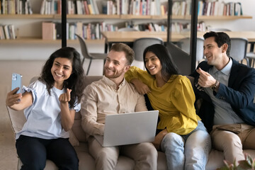 Cheerful Indian woman and group of multiethnic colleagues looking at smartphone camera while take selfie pictures making playful gestures having fun during break sit on couch in modern coworking space