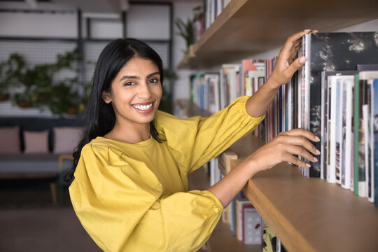 Indian woman standing by bookshelf with lot of diverse literature of university library or bookshop, search information for study or work. Portrait of book lover, hobby, intellectual growth, education