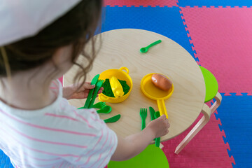 child playing with green toy utensils and food on a small round wooden table, concept of educational games, early childhood education, toy stores, kindergarten