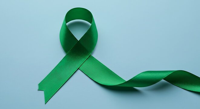 Green awareness ribbon on a light blue background, symbolizing support for mental health, organ donation, and environmental causes with copy space.