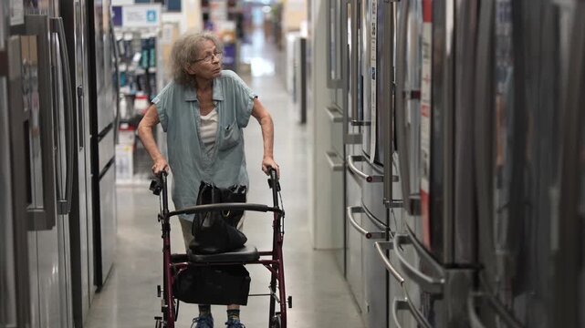 Elderly woman uses a walker while shopping in a hardware store for materials to remodel her home, browsing between rows of appliances and supplies.