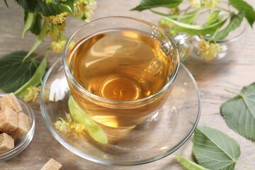 Tasty linden tea in cup, sugar cubes, leaves and flowers on wooden table, closeup