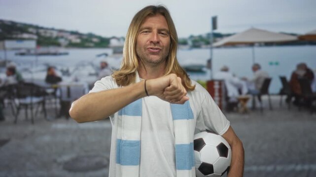 Man with long hair holds ball with arm extended in a fistbump gesture on street wearing fan scarf; enthusiasm.