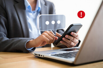 A person in a suit using a smartphone with a message notification and a laptop on the desk nearby