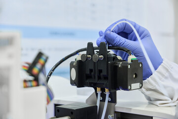Close-up of a blue-gloved hand loading white plastic filament into the injection head of a 3D...