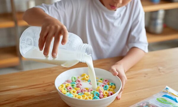 An Asian 12 years old boy wearing a cute t-shirt with is pouring milk from a plastic gallon jug into a white bowl filled with colorful Froot Loops cereal