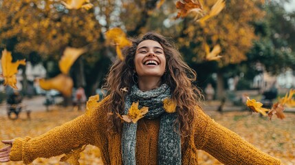 A young woman with curly brown hair smiles joyfully while standing in a park surrounded by autumn leaves. She wears a cozy yellow sweater and a gray scarf.