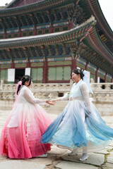 Fototapeta premium In Seoul in June 2025, two Korean women in their twenties wearing pink and sky-blue hanbok stand in front of the traditional brick gate of Gyeongbokgung Palace, their poise echoing ancient grace.