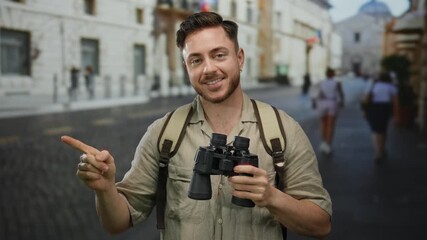 Young man with beard and binoculars smiles on city street pointing while wearing backpack conveying tourist adventure setting on a sunny day outdoors - Powered by Adobe