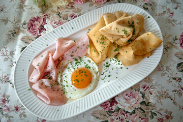 An exquisitely presented breakfast plate featuring soft crepes, a perfectly cooked sunny-side-up egg, and slices of ham garnished with fresh herbs.