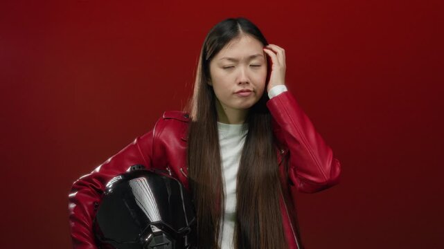 Woman wearing red jacket holding helmet in front of red background appearing thoughtful and focused